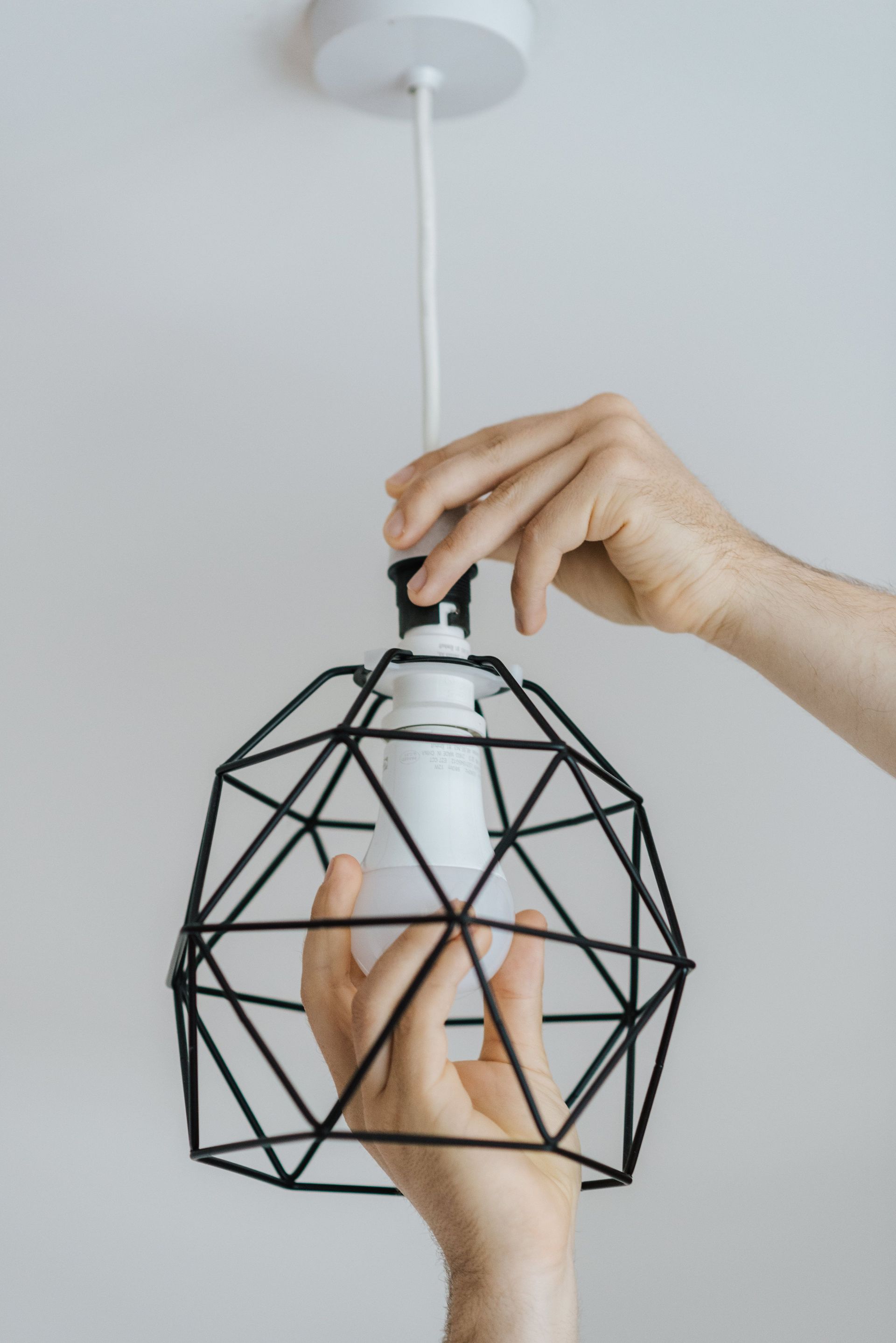 Crop unrecognizable male screwing light bulb into geometric lamp frame hanging on wire in house