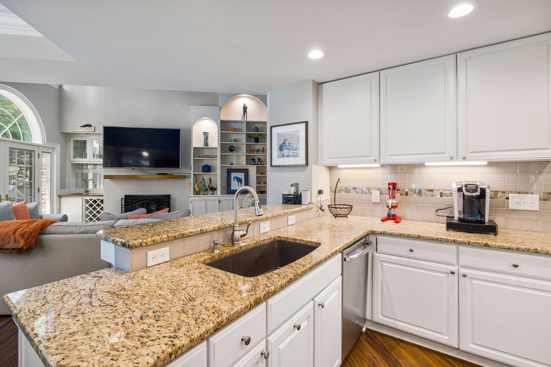 A modern kitchen with granite countertops opens into a stylish living room, featuring elegant furniture and built-in shelves.